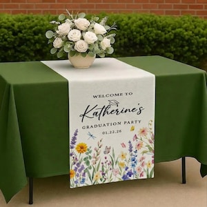 May include: A table set for a graduation party, featuring a green tablecloth and a floral runner. The runner reads "Welcome to Katherine's Graduation Party" with a floral design. A vase of white roses sits atop the table.