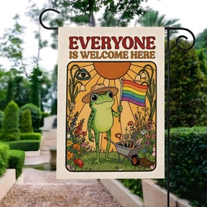 May include: A garden flag with the text "EVERYONE IS WELCOME HERE". The flag features a cartoon frog wearing a hat and holding a rainbow flag. The frog stands in a garden with flowers, a wheelbarrow, and gardening tools. The background is a sunny orange.