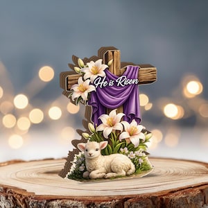 May include: A wooden cross with a purple drape and the text "He is Risen". The cross is decorated with white lilies and a lamb. The lamb is lying down in a bed of flowers and grass.