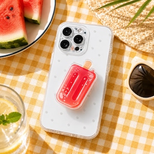 May include: A clear smartphone case featuring a white polka dot pattern and a red popsicle-shaped phone grip attached to the center. The camera module area is surrounded by small star and bow-shaped accents, providing a playful aesthetic.
