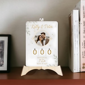 Puede incluir: Un calendario de cuenta regresiva de boda blanco con una foto de una pareja, los nombres "Kelly & Peter" y el texto "Days Until I Do March 16, 2025". El calendario tiene un soporte de madera y detalles florales decorativos.