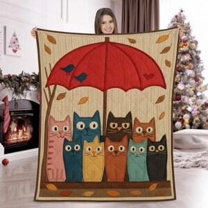 Könnte beinhalten: Eine beige Steppdecke mit einem skurrilen Design von bunten Katzen unter einem roten Regenschirm. Der Regenschirm ist mit zwei blauen Vögeln und Herbstblättern verziert. Die Steppdecke hat einen braunen Rand und ein vertikales Streifenmuster.