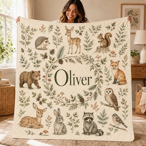 May include: A cream-colored quilted blanket featuring a central wreath of green foliage and the name Oliver in dark green text. The design includes illustrations of a hedgehog, deer, squirrel, bear, fox, owl, rabbit, raccoon, and bird.