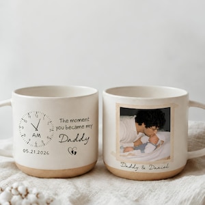 Op de afbeelding: Twee gebroken witte keramische mokken met een gespikkelde afwerking. Eén mok heeft een klokafbeelding op AM, de tekst "The moment you became my Daddy", de datum "05.21.2026" en een hartje met voetafdrukken. De andere mok toont een foto en "Daddy & Daniel".