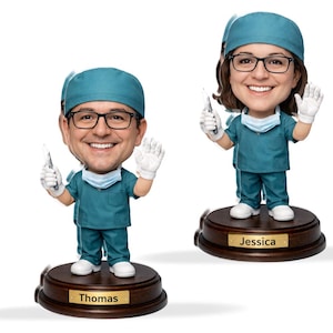 Op de afbeelding: Twee bobblehead-beeldjes van medische professionals. Elke figuur draagt ​​een tealkleurige operatiejas, een chirurgische muts, een bril en een gezichtsmasker. Eén figuur is gelabeld met "Thomas" en de andere met "Jessica". Beide figuren houden een chirurgisch instrument vast en zwaaien.