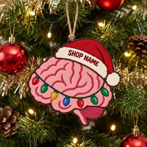 Può includere: Un ornamento a forma di cervello rosa con un cappello da Babbo Natale rosso e bianco con il testo SHOP NAME stampato sul bordo. Il cervello è avvolto da una serie di luci natalizie rosse, verdi, gialle e blu, appeso a un cordino di juta.