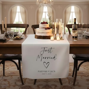 Peut inclure: Une table de réception de mariage avec un chemin de table blanc sur lequel on peut lire "Just Married" avec un cœur, des noms et une date. Un gâteau à deux étages avec des baies repose sur une base en bois. Des bougies, des verres à vin et une bouteille de vin sont également sur la table.
