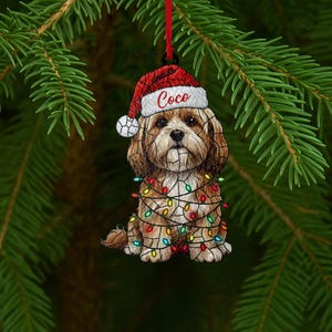 Peut inclure: Ornement de Noël représentant un chien marron et blanc portant un bonnet de Père Noël avec le nom "Coco" et enveloppé de guirlandes lumineuses colorées. L'ornement est suspendu à un ruban rouge, sur fond d'aiguilles de pin vertes.