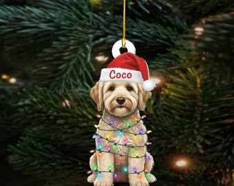 May include: A Christmas ornament featuring a light brown dog wearing a Santa hat with the name "Coco" and wrapped in colorful Christmas lights. The ornament hangs from a gold string against a backdrop of green Christmas tree branches.