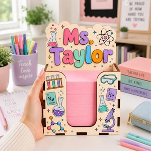 May include: A wooden desk organizer featuring the name Ms. Taylor in colorful letters, decorated with science-themed illustrations including beakers, a microscope, and chemical elements. The front holds a stack of pink sticky notes.