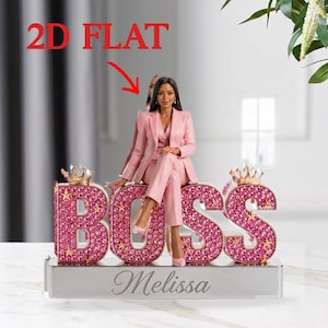 Könnte beinhalten: Eine rosa Figur einer Frau in einem Anzug, die auf dem Wort "BOSS" sitzt, das mit rosa Edelsteinen geschrieben ist. Die Figur steht auf einer silbernen Basis mit dem eingravierten Namen "Melissa". Zwei goldene Kronen befinden sich auf den Buchstaben.