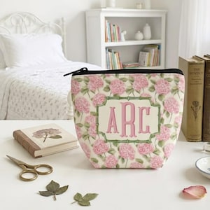 Puede incluir: Un neceser con estampado floral y cremallera negra, con flores de hortensia rosas sobre un fondo crema. La bolsa tiene un marco rectangular con las letras "ARC" en rosa. Un cuaderno, tijeras y hojas secas están sobre la mesa.