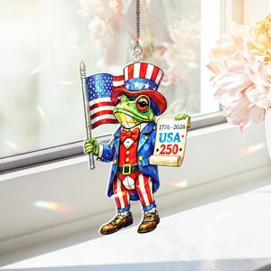 Puede incluir: Un adorno de rana patriótica, vestida con un atuendo del Tío Sam rojo, blanco y azul, sostiene una bandera estadounidense y un pergamino con "USA 250". La rana lleva un sombrero de copa y una pajarita. El adorno está diseñado para colgar.