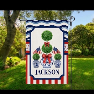 May include: A garden flag with a patriotic design. The flag features topiary trees with red bows, set in blue and white vases. American flags flank the trees. The name "JACKSON" is displayed at the bottom, with a red, white, and blue border.