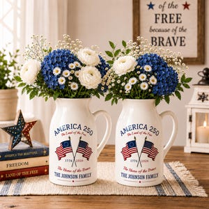 May include: Two white ceramic pitchers with handles, each adorned with the text "AMERICA 250" and American flags. They hold bouquets of blue hydrangeas, white daisies, and other flowers. The text "The Home of the Brave" and "THE JOHNSON FAMILY" are also printed on the pitchers.