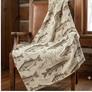 Puede incluir: Una manta beige con un estampado de peces, colocada sobre una silla de madera. La manta presenta ilustraciones de diferentes peces en tonos marrones. La silla tiene un asiento y respaldo de cuero marrón, con un marco de madera.