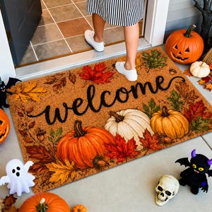 May include: A rectangular coir doormat featuring the word welcome in black cursive script, surrounded by a printed pattern of orange and white pumpkins, red and yellow autumn leaves, and brown acorns on a natural tan textured background.