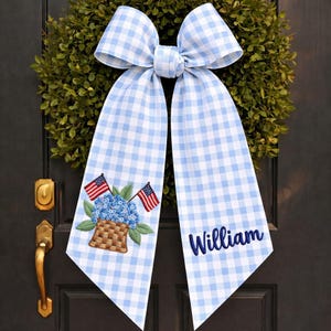 May include: A light blue and white gingham bow with embroidered details. The bow features a basket of blue flowers with American flags and the name "William" in navy blue script. The bow is attached to a green wreath.