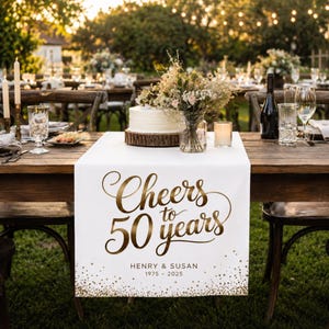 Può includere: Una tovaglia bianca con testo dorato che recita "Cheers to 50 years" e i nomi "Henry & Susan" con gli anni "1975-2025". La tovaglia è su un tavolo di legno apparecchiato per una festa, con una torta, fiori e candele.
