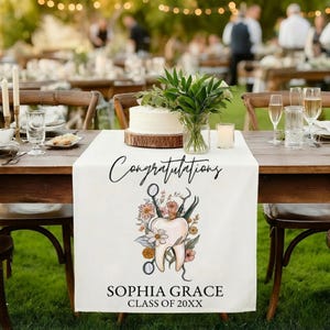 May include: White table runner with the word "Congratulations" in black script. A tooth graphic with dental tools and flowers is below the word. The name "SOPHIA GRACE" and "CLASS OF 20XX" are printed below the graphic.