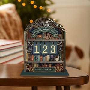 May include: A decorative wooden bookshelf-shaped calendar with the text "BOOKS READ THIS YEAR" and the numbers "123" displayed. The calendar features a dragon and the words "ASHLEY'S LIBRARY". The design includes miniature books and a small clock.