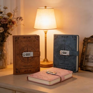 Könnte beinhalten: Drei ledergebundene Journale mit Zahlenschlössern. Eines ist braun mit den Texten "YOUR NAME" und "TROUVAILLE", ein anderes ist blau mit den Texten "Ken" und "TROUVAILLE", und das dritte ist rosa. Ein Kompass-Design befindet sich auf den Einbänden.