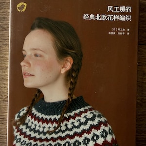 Könnte beinhalten: Buchcover mit einer Frau in einem gemusterten Pullover. Das Cover ist braun mit japanischem Text und einem goldenen Emblem. Der Pullover hat ein weißes, rotes und schwarzes geometrisches Design. Das Buch handelt vom Stricken.