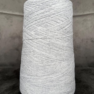 Puede incluir: Una gran bobina cilíndrica de hilo gris claro. El hilo está enrollado firmemente, creando una superficie texturizada. El color es un gris suave y neutro, adecuado para varios proyectos de tejido o ganchillo. El hilo está sobre una superficie oscura.