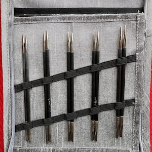 Peut inclure: Un étui en tissu gris avec une fermeture éclair contenant plusieurs aiguilles à tricoter noires avec des pointes argentées. Les aiguilles sont maintenues par des bandes élastiques noires. L'étui est sur une surface rouge.