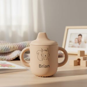 May include: A beige sippy cup with a lid and two handles. A cartoon squirrel is printed on the front, with the name "Brian" below. The cup is made of a soft material and is designed for babies and toddlers.