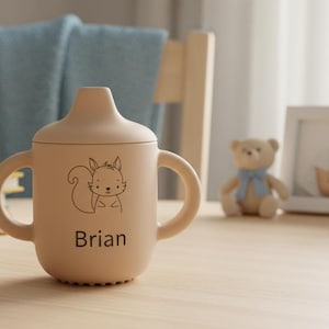 May include: A tan sippy cup with a squirrel graphic and the name "Brian" printed on it. The cup has two handles and a lid. A teddy bear and framed photos are in the background.