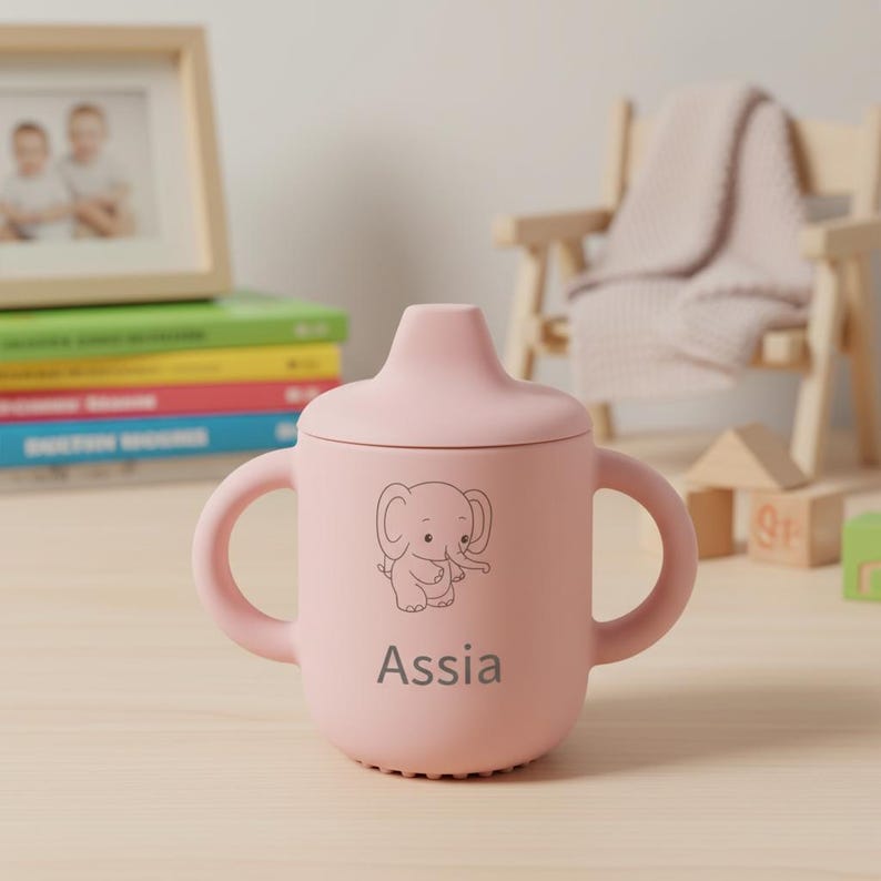 May include: A pink sippy cup with a lid and two handles. The cup features a cartoon elephant illustration and the name "Assia" printed below the elephant. The cup is sitting on a light-colored wooden surface.