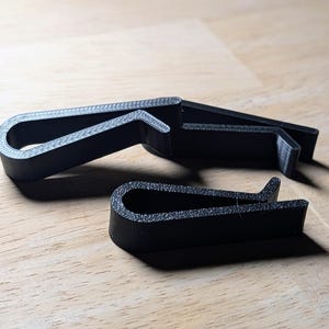 May include: Three black plastic clips are arranged on a light wood surface. The clips have a curved, spring-like design with a textured surface. The clips are likely used for holding items together.