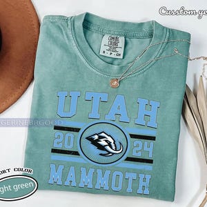 May include: A light green t-shirt with the words "UTAH MAMMOTH" in light blue. The shirt features a graphic of a mammoth and the numbers "20 24". The shirt is labeled "COMFORT COLORS" and "light green". A silver necklace is draped across the shirt.