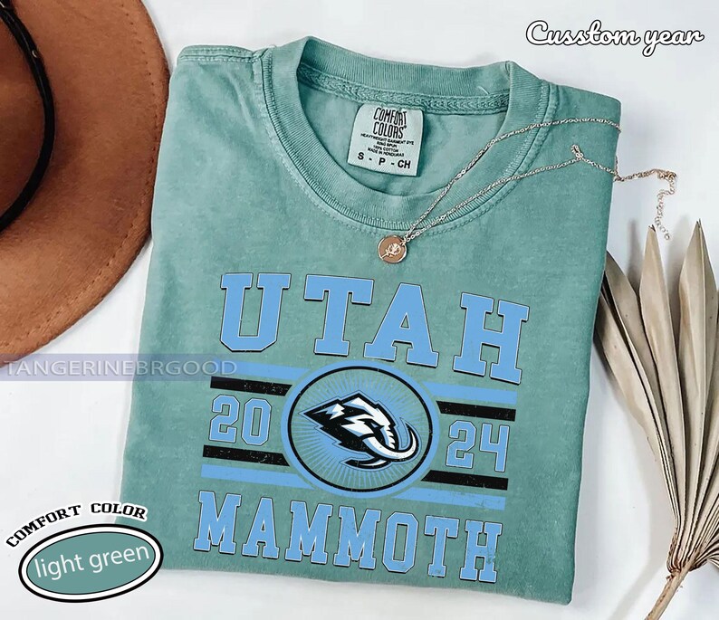 May include: A light green t-shirt with the words "UTAH MAMMOTH" in light blue. The shirt features a graphic of a mammoth and the numbers "20 24". The shirt is labeled "COMFORT COLORS" and "light green". A silver necklace is draped across the shirt.