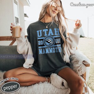 May include: Dark grey t-shirt with the text "UTAH MAMMOTH" in light blue, with a circular emblem featuring a mammoth. The year "2024" is also displayed. The shirt is a comfort color, pepper.