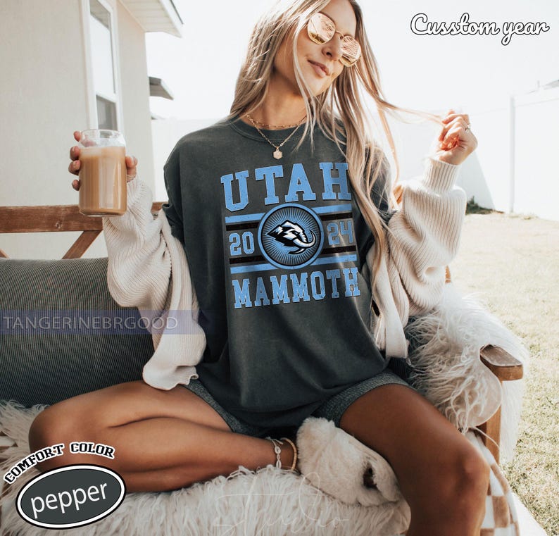 May include: Dark grey t-shirt with the text "UTAH MAMMOTH" in light blue, with a circular emblem featuring a mammoth. The year "2024" is also displayed. The shirt is a comfort color, pepper.