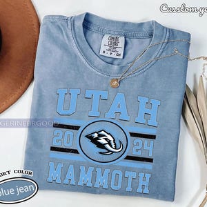 May include: A blue jean Comfort Colors t-shirt with the word "UTAH" in light blue block letters. Below the word "UTAH" is a circular logo with a mammoth graphic. The text "2024" and "MAMMOTH" are also printed on the shirt.