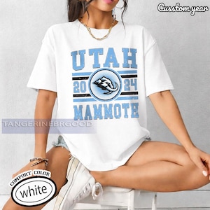 May include: White t-shirt with a light blue graphic that says "UTAH MAMMOTH 2024". The graphic includes a circular emblem with an elephant design. The shirt has a crew neck and short sleeves.