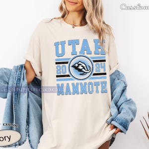 May include: Ivory-colored t-shirt with the text "UTAH 2024 MAMMOTH" in light blue. The design includes a circular emblem with a stylized mammoth graphic. The shirt is part of the "Comfort Color" collection.