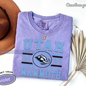 May include: A violet Comfort Colors t-shirt with the text "UTAH" in light blue block letters. Below the text is a circular logo with a mammoth graphic and the numbers "20" and "24". The word "MAMMOTH" is printed below the logo. A gold necklace rests on the shirt.