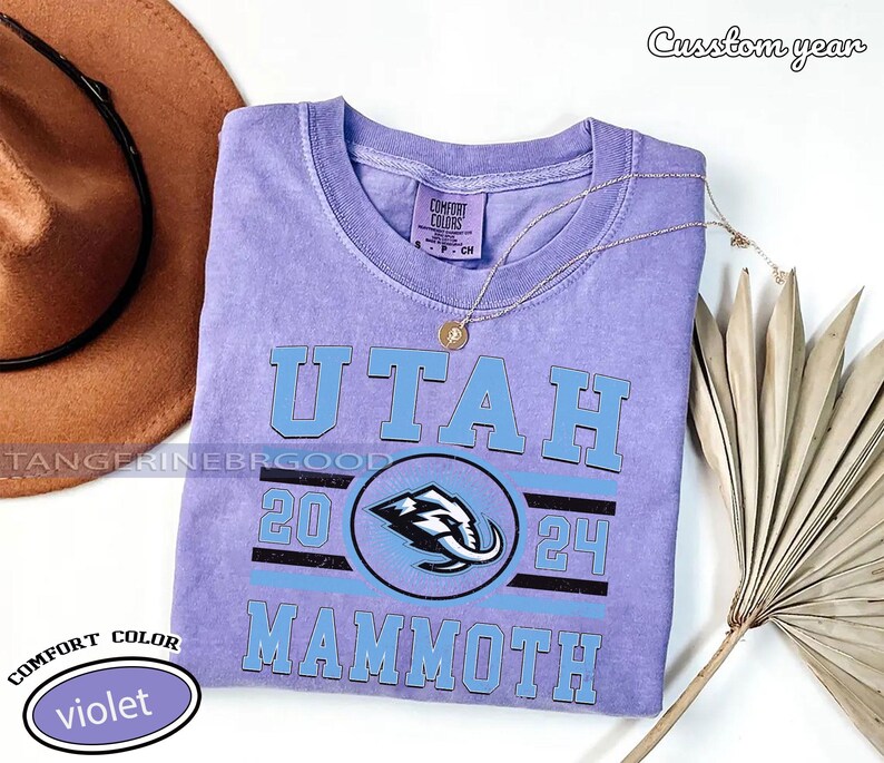 May include: A violet Comfort Colors t-shirt with the text "UTAH" in light blue block letters. Below the text is a circular logo with a mammoth graphic and the numbers "20" and "24". The word "MAMMOTH" is printed below the logo. A gold necklace rests on the shirt.