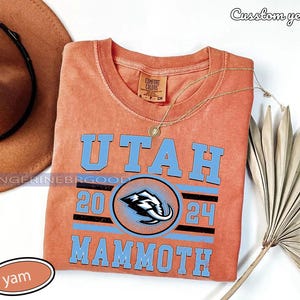 May include: An orange crew neck t-shirt with the word "UTAH" in light blue block letters. Below the word "UTAH" is the number "20" and "24" with a Mammoth logo in the center. A gold necklace and a brown hat are also in the image.
