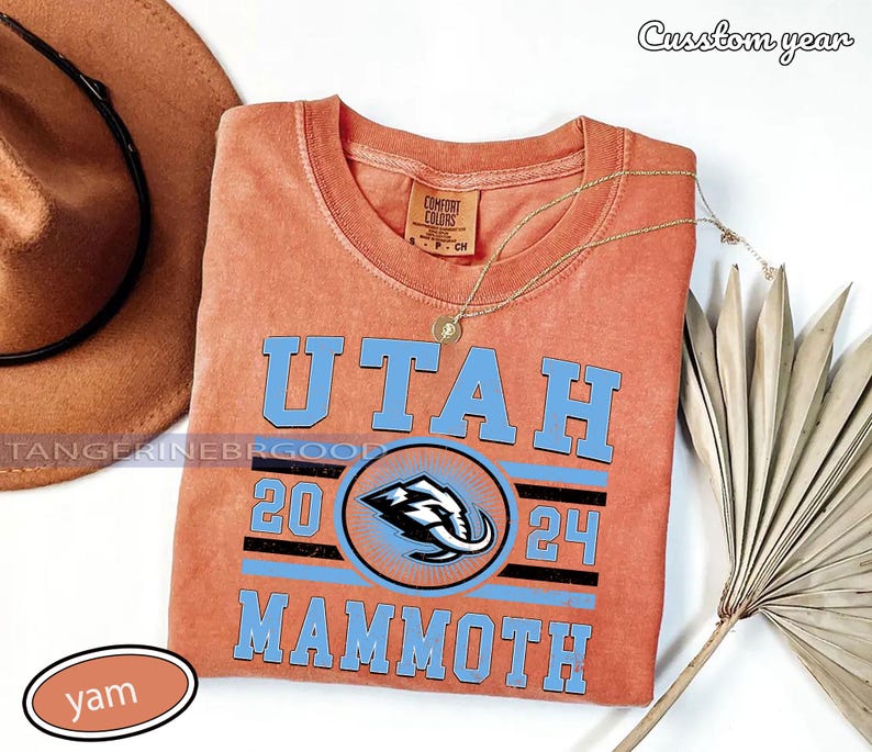 May include: An orange crew neck t-shirt with the word "UTAH" in light blue block letters. Below the word "UTAH" is the number "20" and "24" with a Mammoth logo in the center. A gold necklace and a brown hat are also in the image.