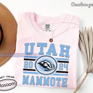 May include: A light pink t-shirt with the text "UTAH MAMMOTH 2024" in light blue. The shirt features a circular logo with a mammoth graphic. A gold necklace with a pendant is draped on the shirt.