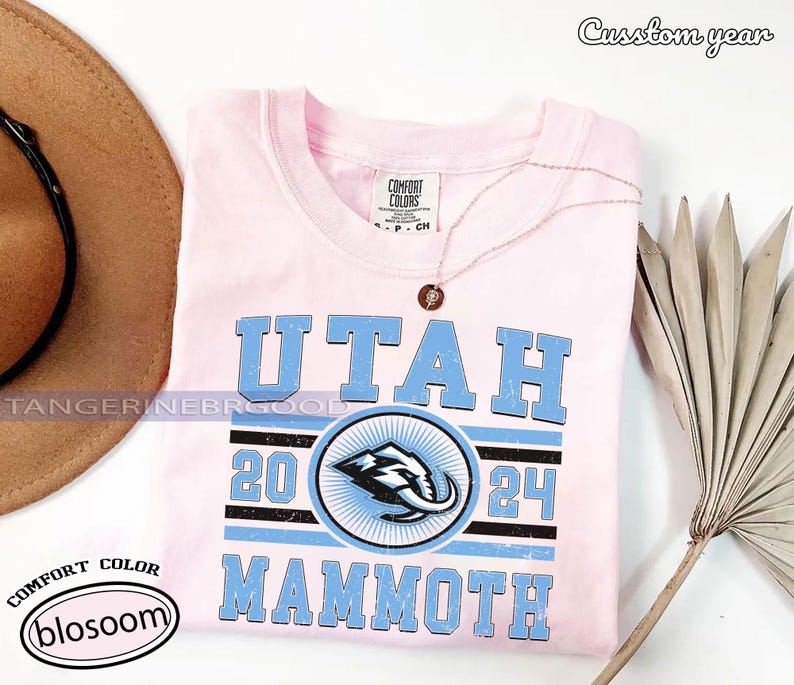 May include: A light pink t-shirt with the text "UTAH MAMMOTH 2024" in light blue. The shirt features a circular logo with a mammoth graphic. A gold necklace with a pendant is draped on the shirt.