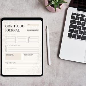Può includere: Un diario di gratitudine digitale visualizzato su un tablet. Il diario ha il titolo "GRATITUDE JOURNAL". Le pagine del diario contengono suggerimenti per le riflessioni quotidiane. Sono visibili anche uno stilo bianco, un laptop e una piccola pianta in vaso.