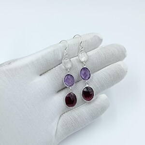 Puede incluir: Pendientes colgantes con tres piedras preciosas ovaladas en tonos claros, morados y rojo oscuro. Las piedras están engastadas en un marco de metal plateado. Los pendientes cuelgan de un gancho plateado.