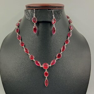May include: A silver necklace and earring set displayed on a black mannequin. The jewellery features oval and teardrop-shaped red gemstones. The necklace has a central pendant with three gemstones. The earrings have a single teardrop-shaped gemstone.