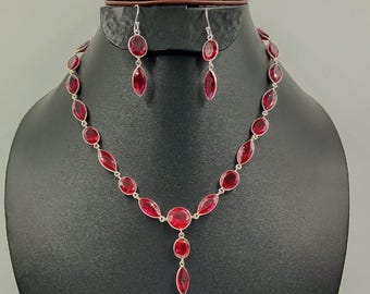 以下が含まれることがあります： 黒いマネキンに展示されたシルバーのネックレスとイヤリングのセット。ジュエリーは、楕円形と涙滴型の赤い宝石が特徴です。ネックレスには、3つの宝石が付いた中央のペンダントが付いています。イヤリングには、1つの涙滴型の宝石が付いています。